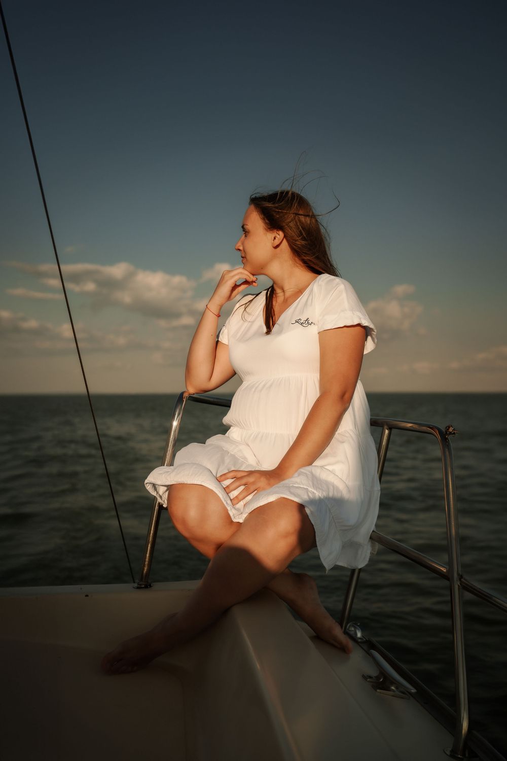 Woman in summer dress sits thoughtfully on yacht deck during sunset cruise.