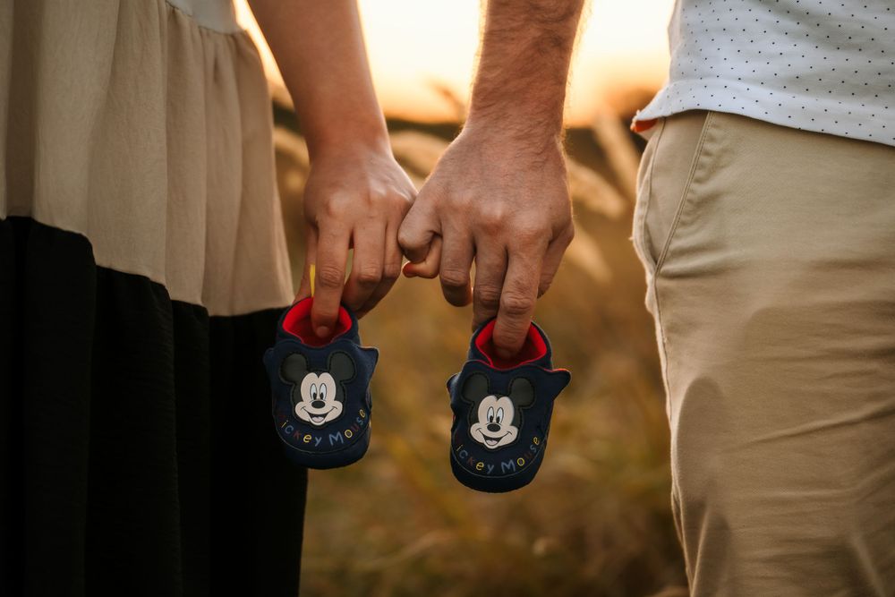 Close-up of hands holding Mickey Mouse baby shoes during sunset pregnancy announcement.
