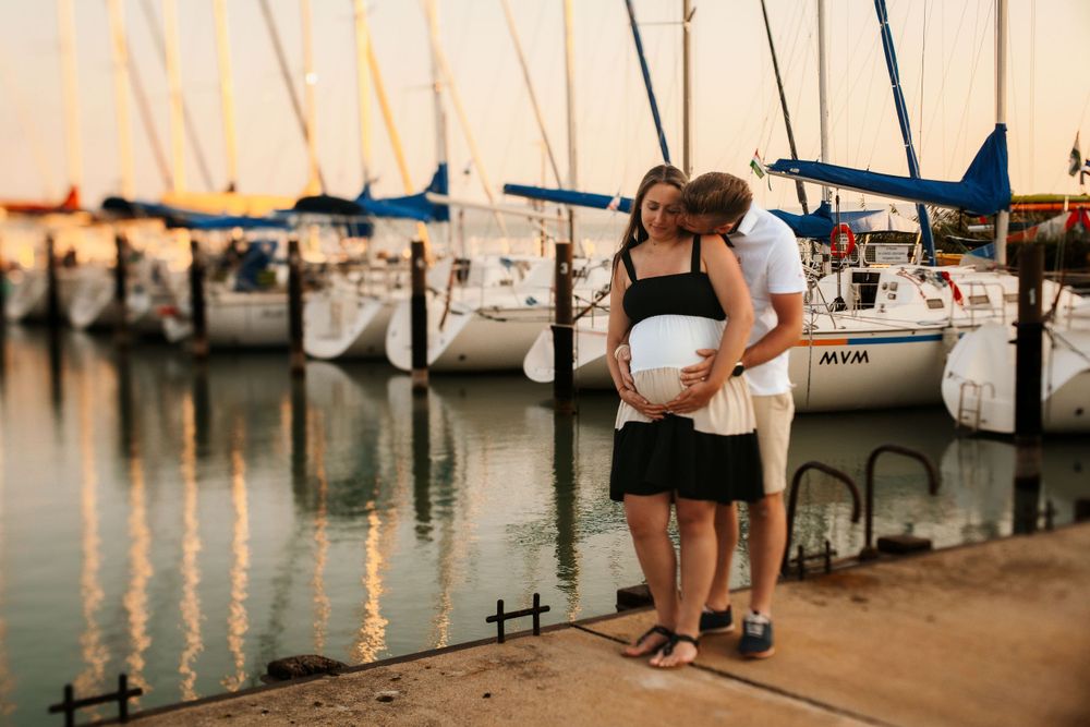 A couple embraces while watching sailboats at a marina during golden hour sunset.