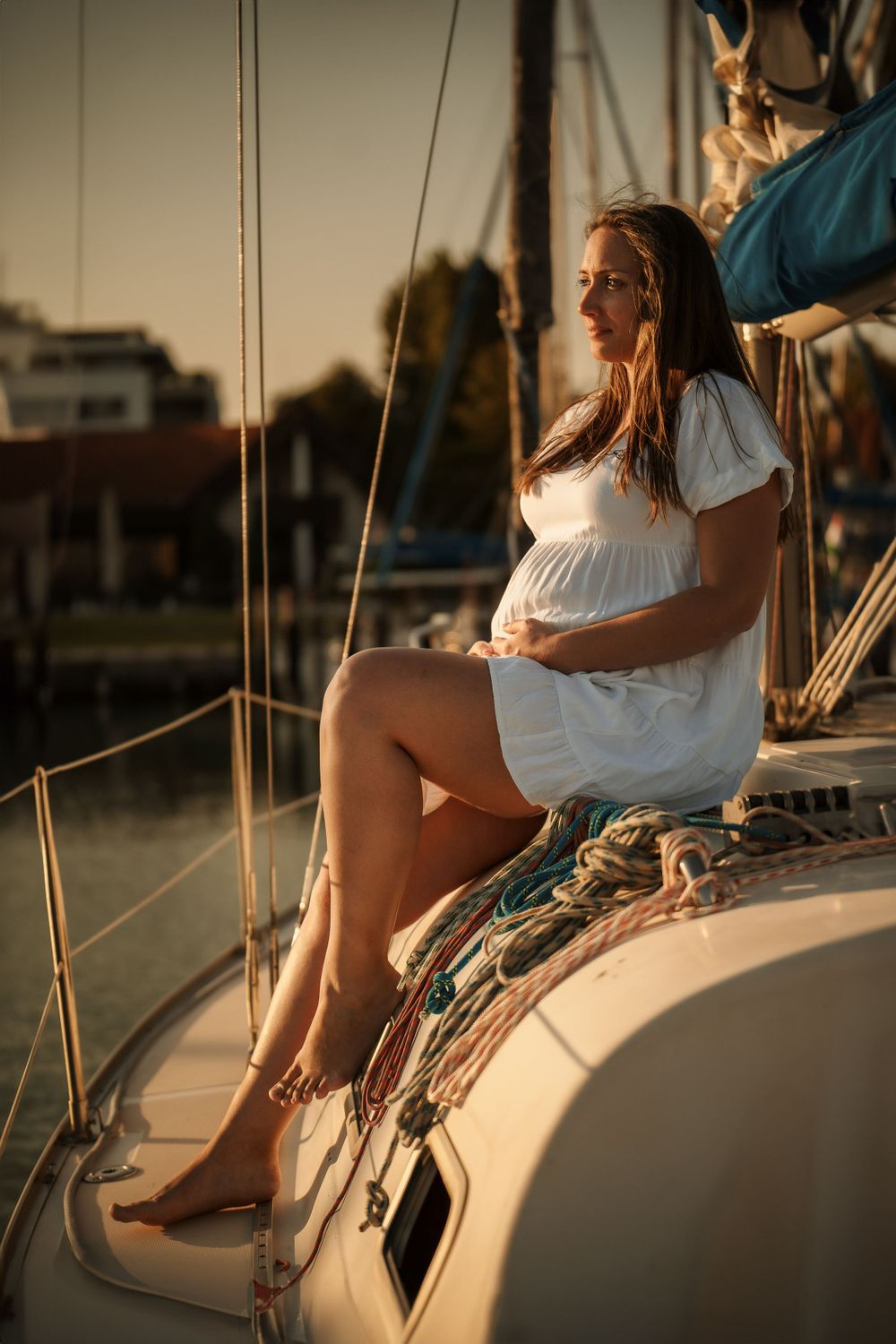 A maternity photo on a sailboat at sunset features a woman in a white dress.