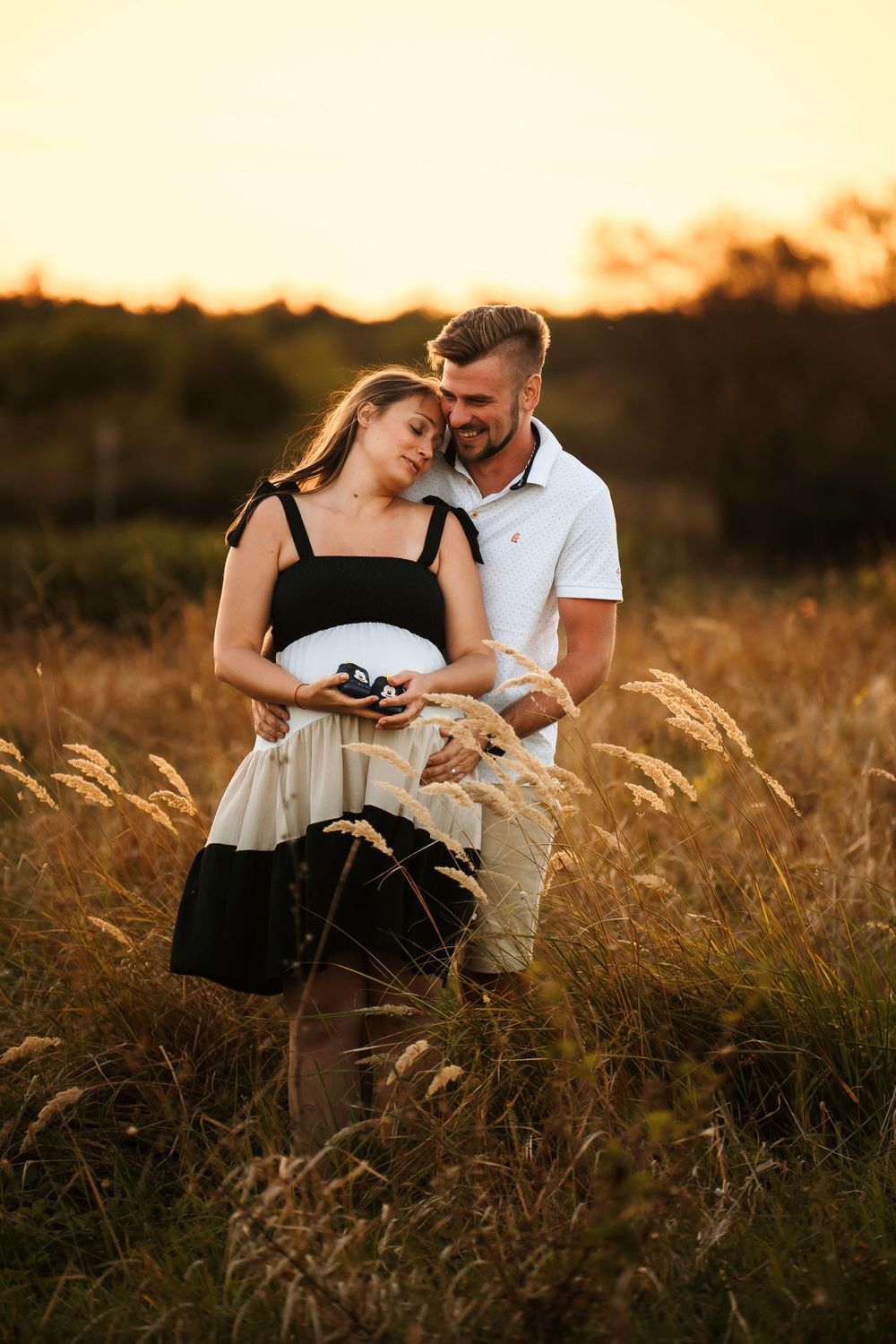Tender moment between couple holding baby shoes in wheat field at sunset.