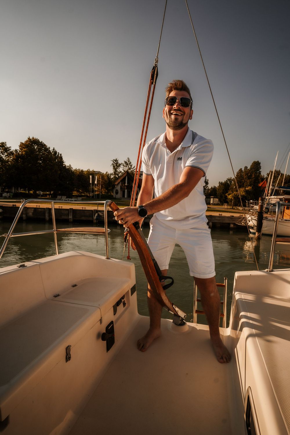 Person in white summer attire standing on a boat deck holding sailing equipment.