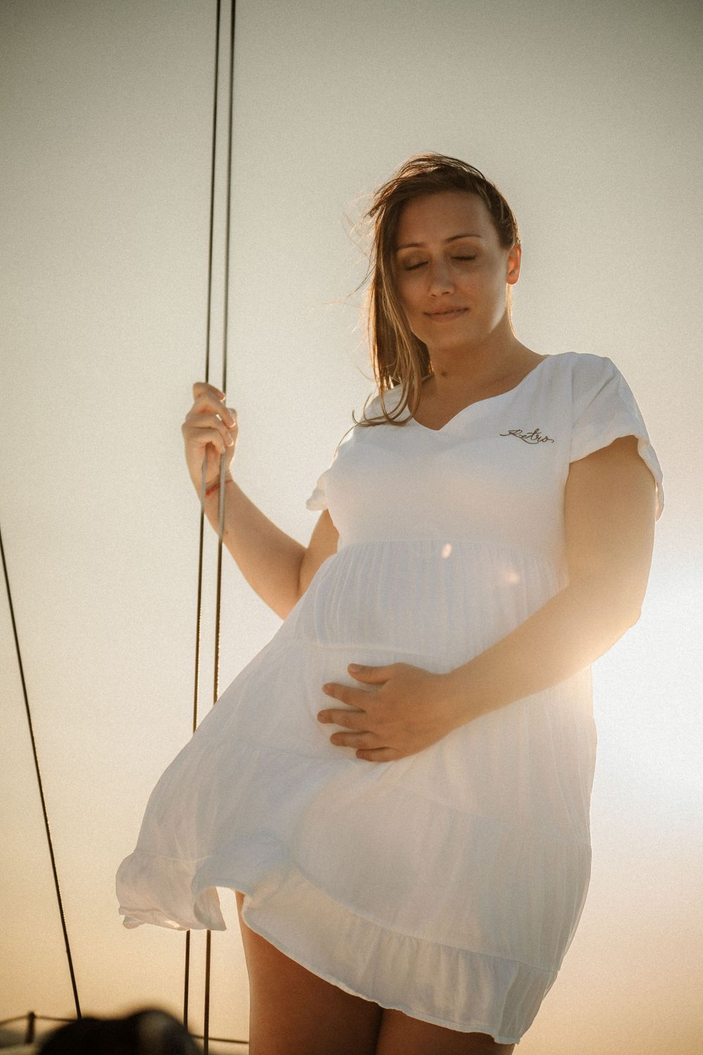 Woman in white dress poses on sailboat deck during golden hour with wind in her hair.