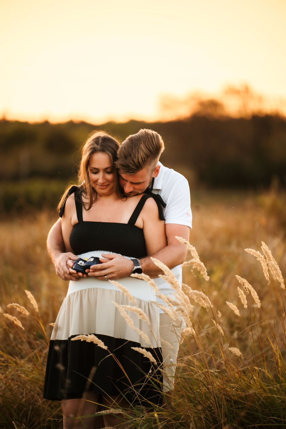 Couple embracing while looking at baby shoes during sunset pregnancy announcement photoshoot.
