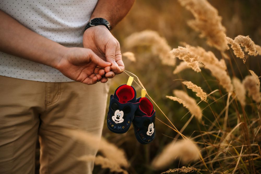 Close-up of hands holding small Mickey Mouse themed baby shoes against golden wheat stalks.