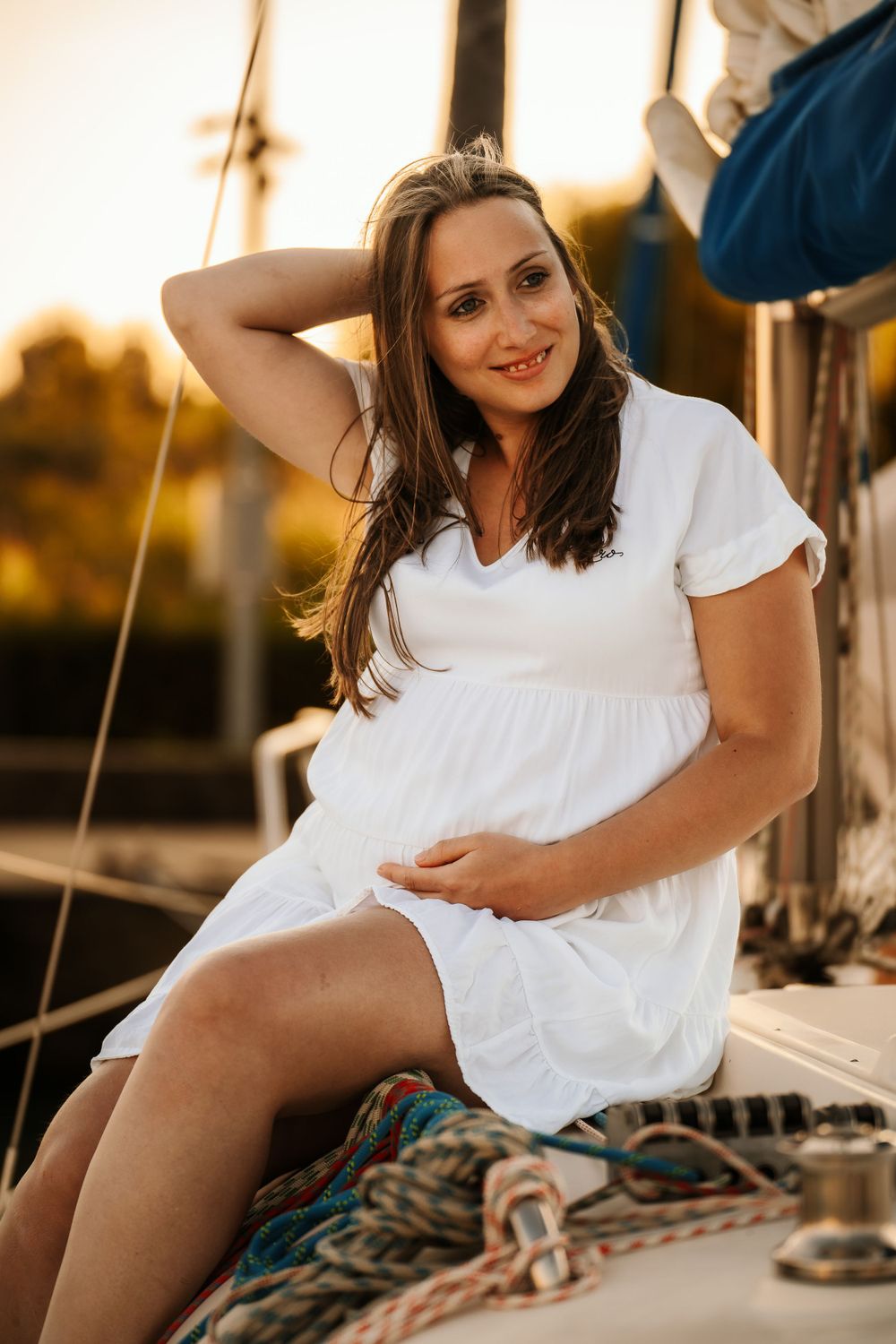 Woman in white summer dress posing on sailboat deck during golden sunset.