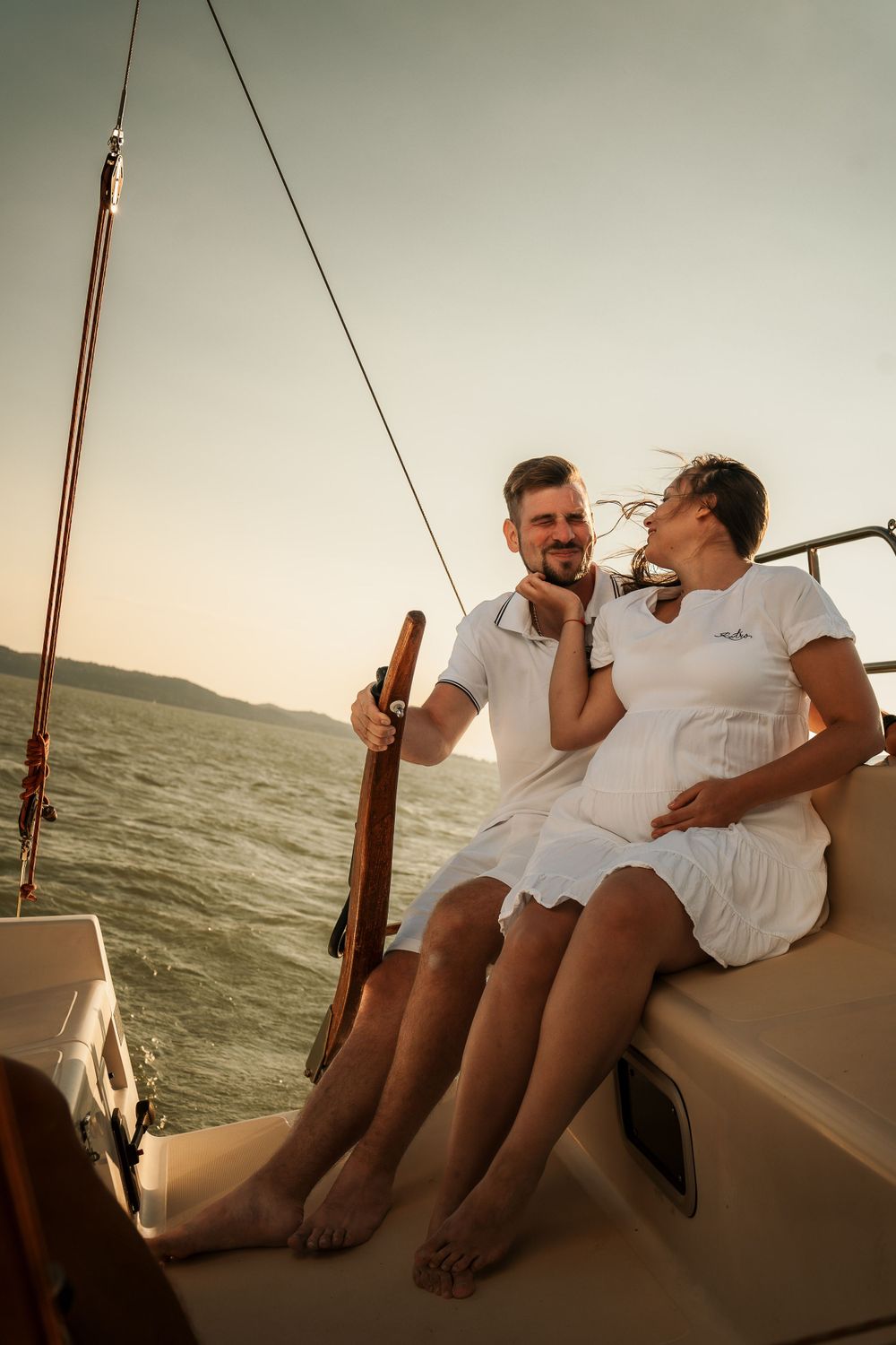 Couple in white outfits sharing an intimate moment on a sailboat at sunset.