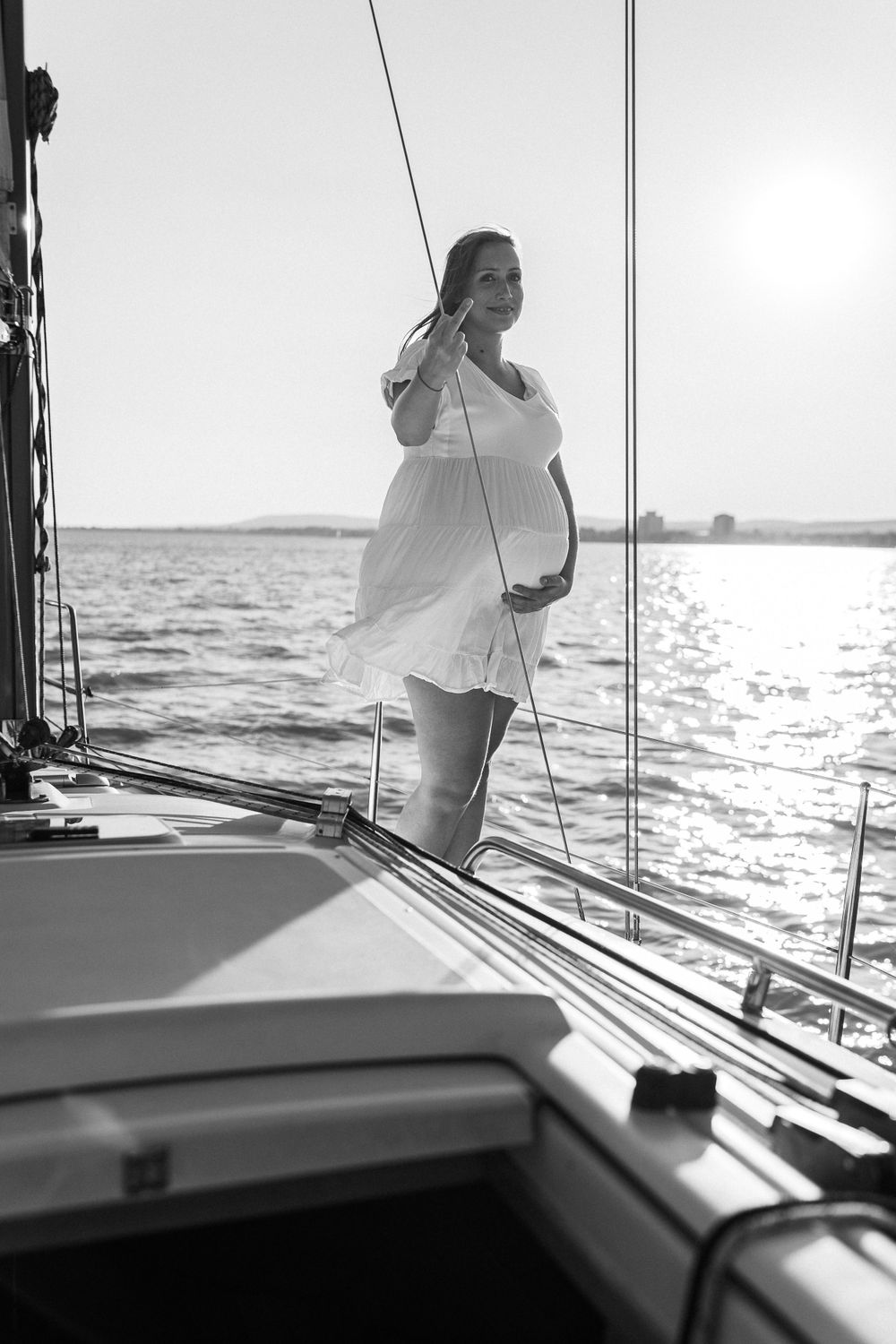 Woman in flowing white dress stands on sailboat deck during sunset over calm waters.