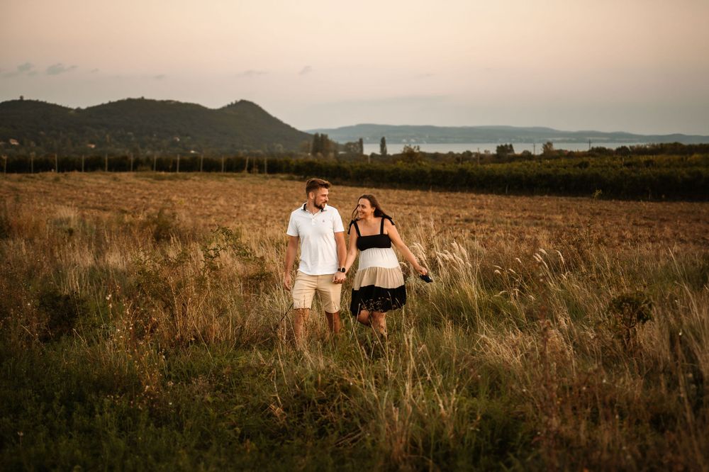 Couple stands in field with mountains and lake in background during sunset.