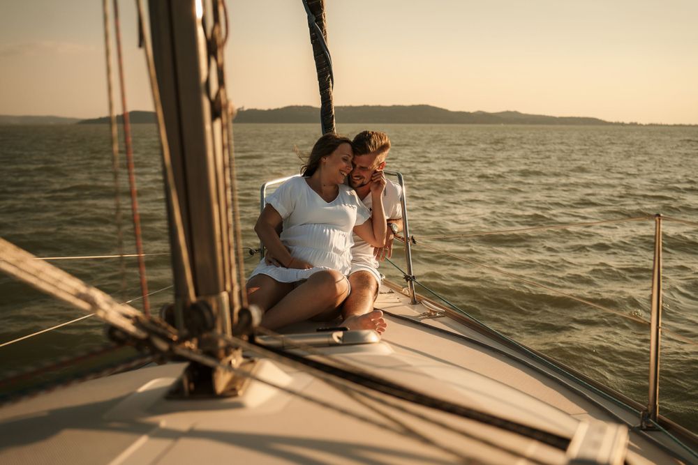 Two people relaxing on a sailboat deck while cruising on calm waters at sunset.