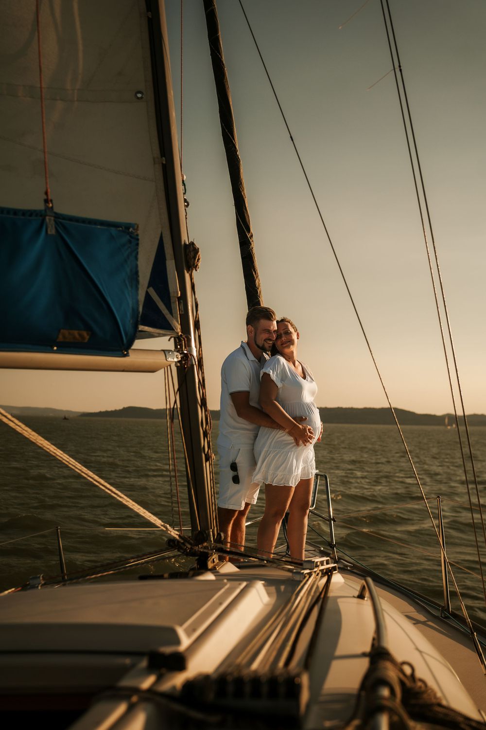 Couple in white clothing embracing on the deck of a sailboat during sunset.