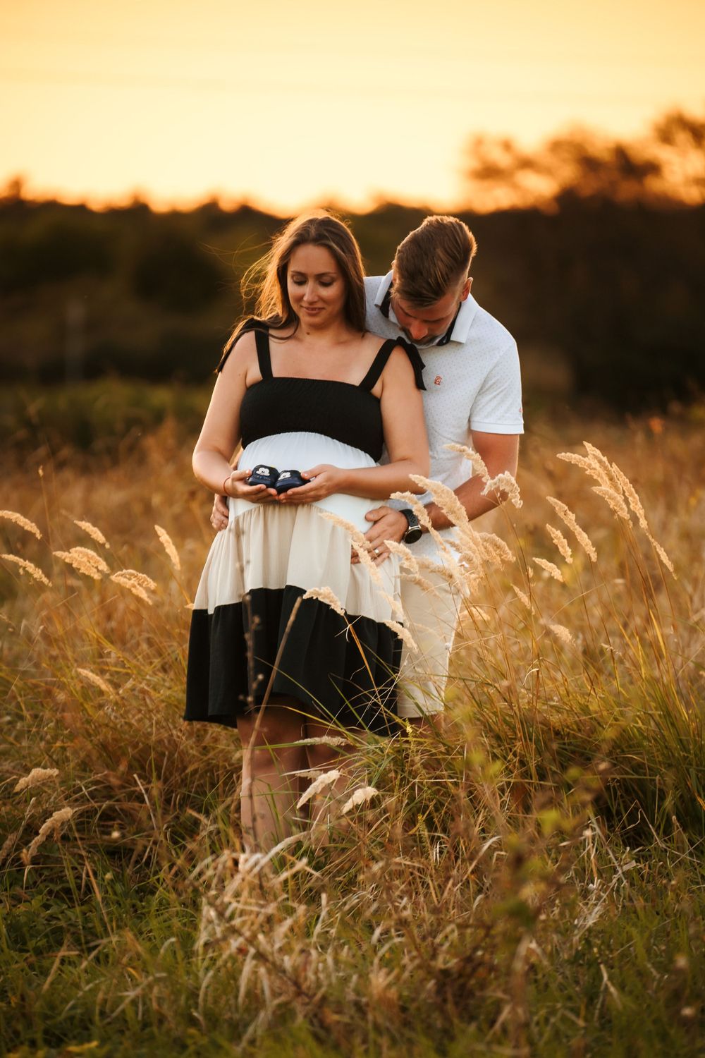 Couple sharing intimate moment while holding baby shoes in golden sunset field.