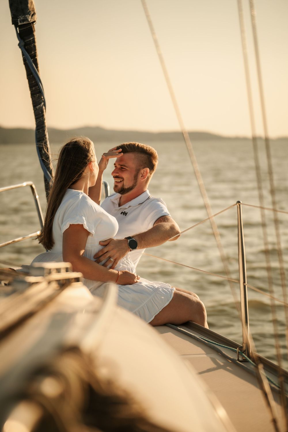 Couple shares an intimate moment on a sailboat at sunset with water and mountains in background.