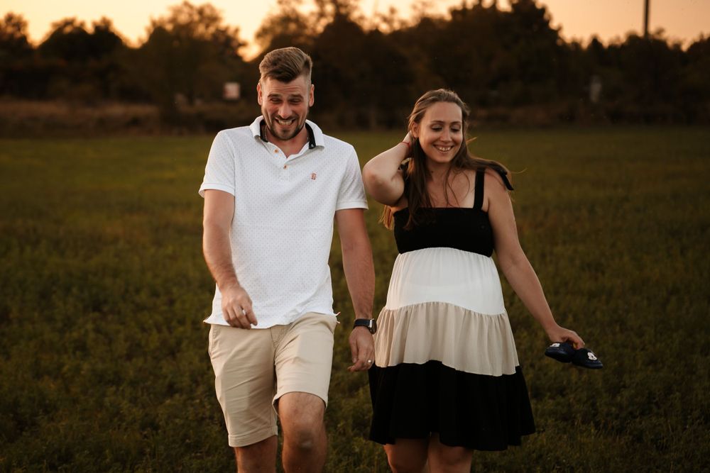 A couple walks together through a grassy field at sunset during a maternity photoshoot.