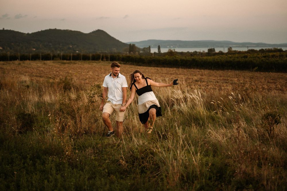 Couple walks through golden field with mountains and water in background.