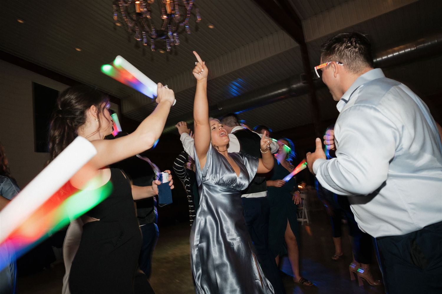 Wedding guests dance and wave colorful glow sticks during evening reception celebration.