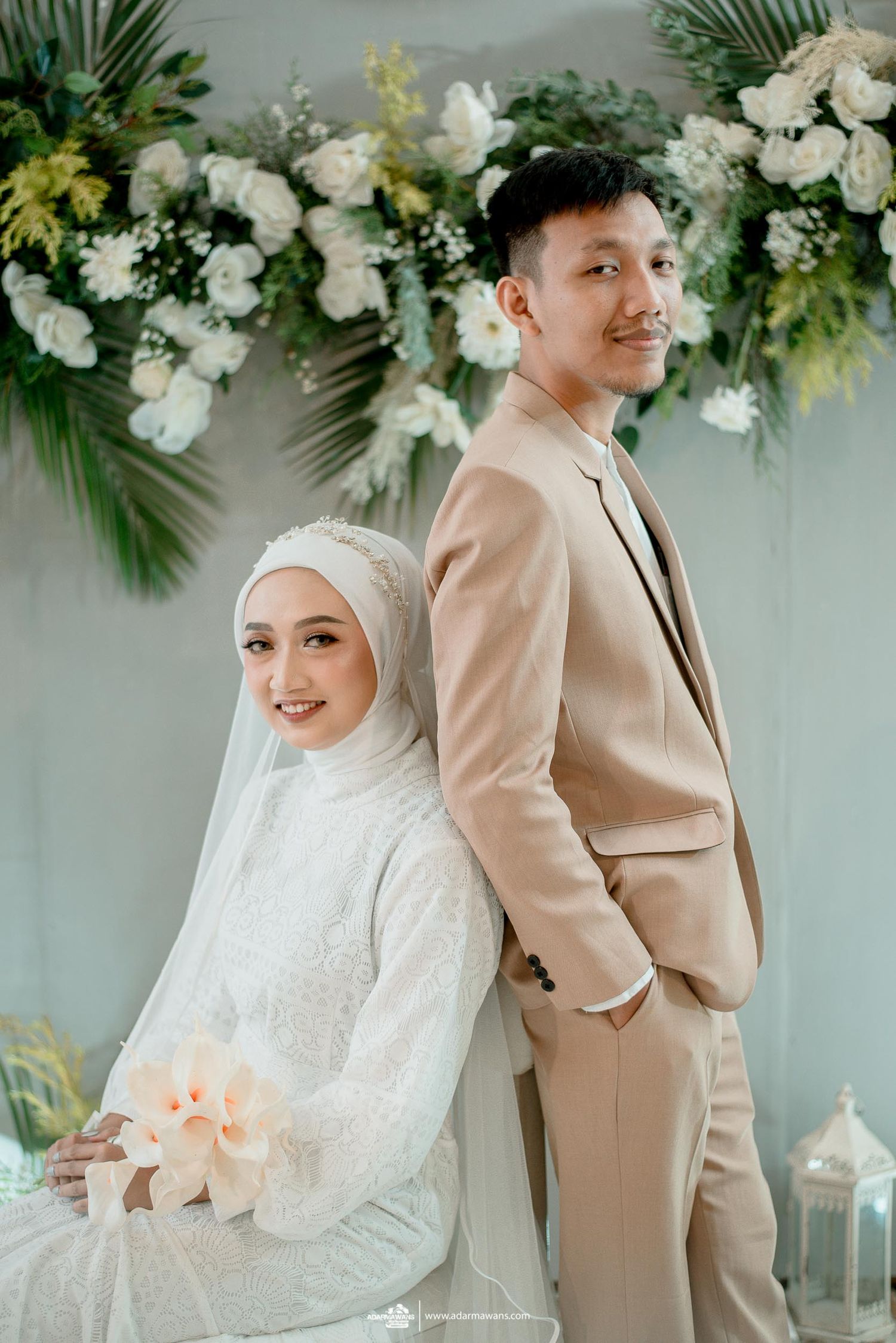 Muslim couple pose against floral backdrop in beige suit and white wedding dress with hijab.