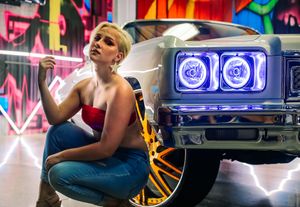 blonde woman in jeans and red tube top crouched next to a vintage muscle car with purple headlights in front of neon lights and graffiti