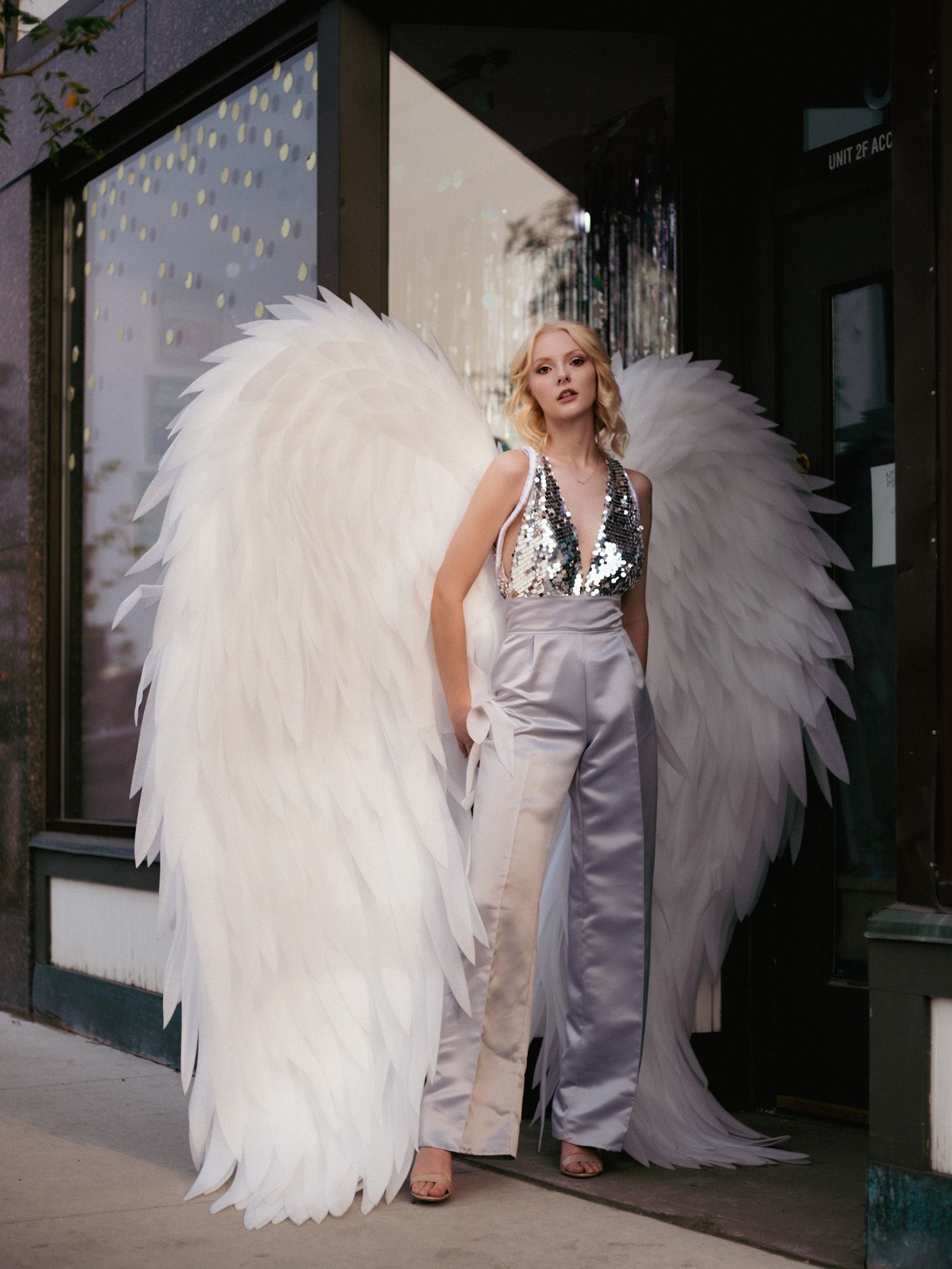 blonde woman in silver jumpsuit stands in an urban area with large angel wings behind her