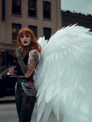 woman with red hair and lots of tattoos wearing a black tanktop, jeans, and large angel wings behind her
