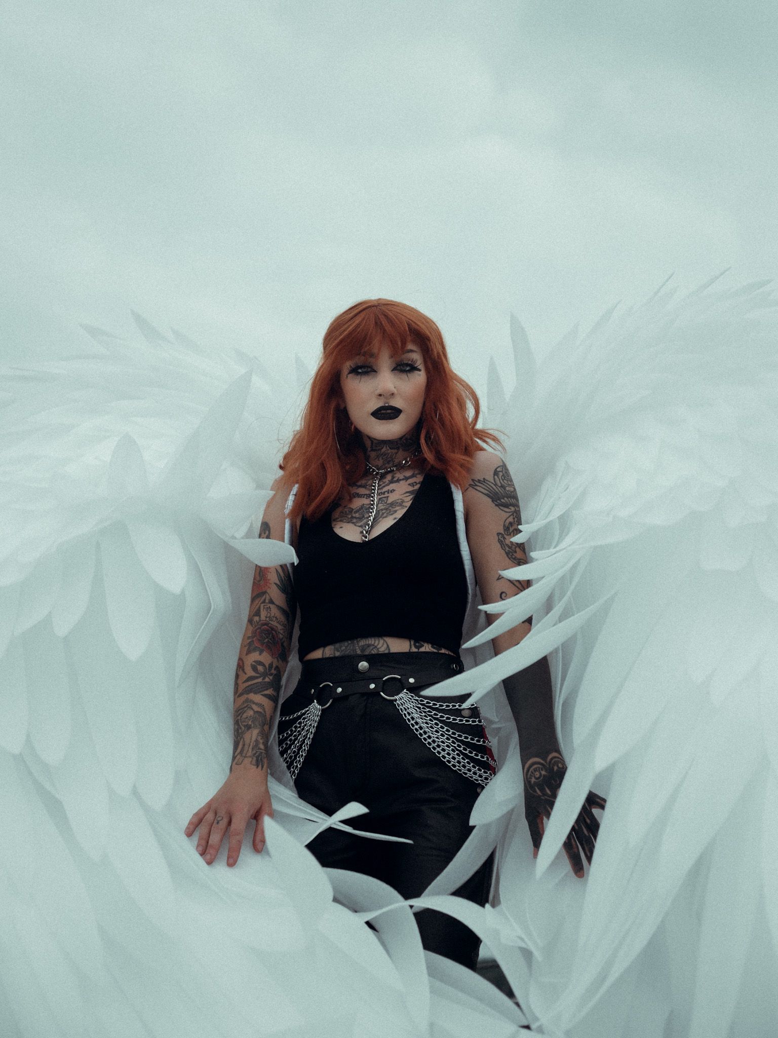 woman with red hair and lots of tattoos wearing a black tanktop, jeans, and large angel wings surrounding her