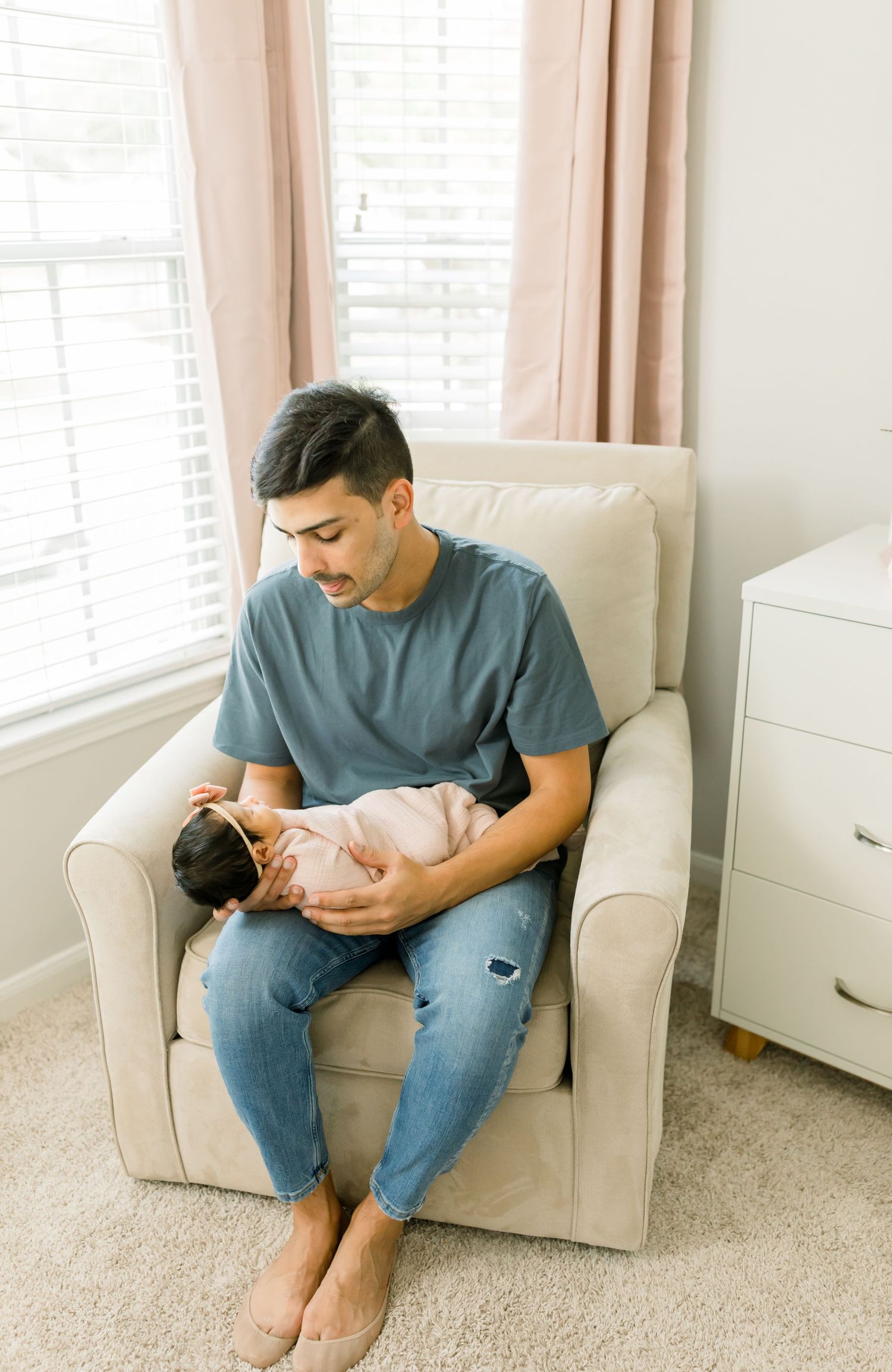 Parent tenderly holds sleeping newborn baby in a cozy beige armchair by a sunny window.