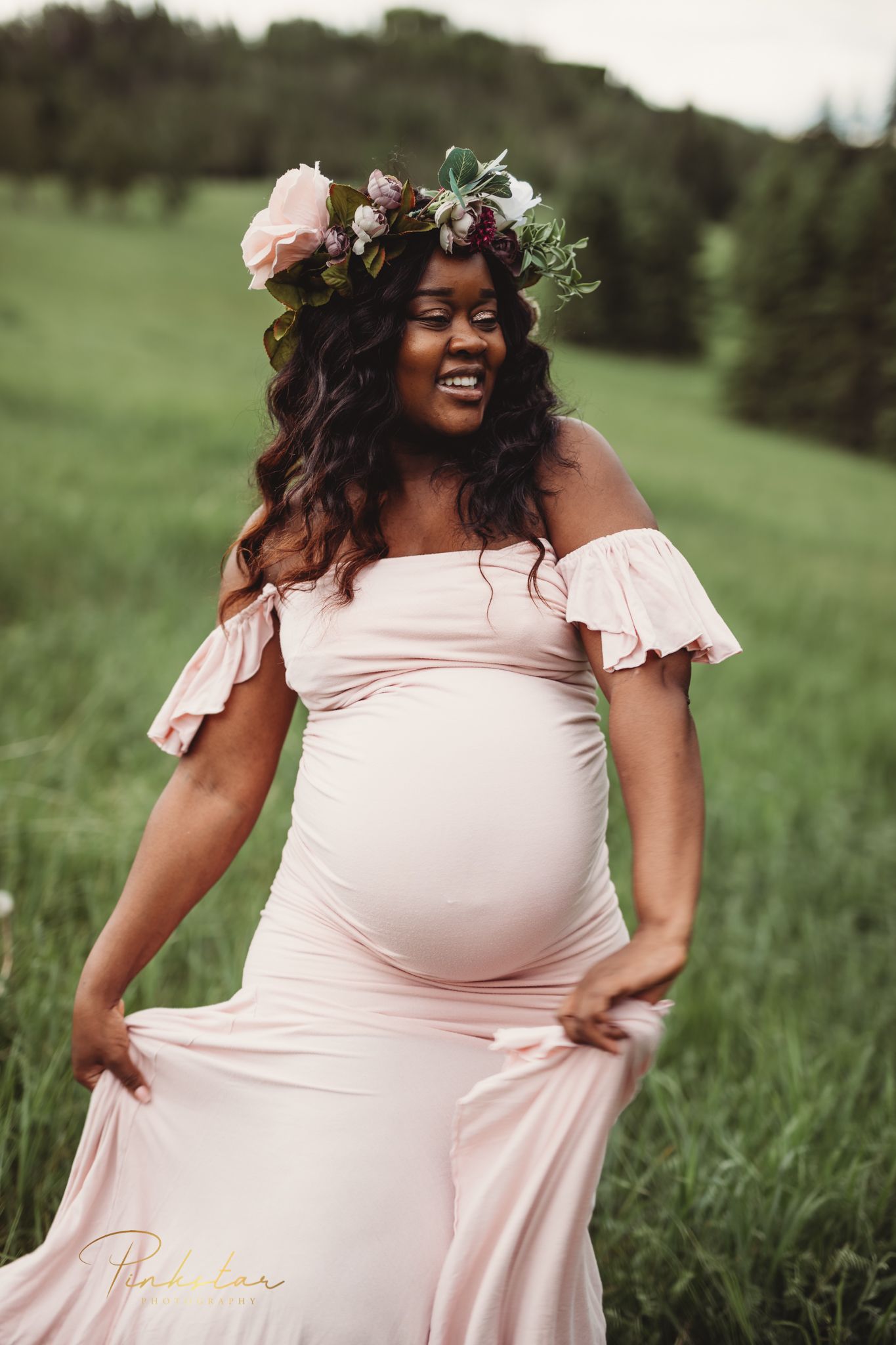 Expecting mother in pink off-shoulder dress and floral crown poses in green field.