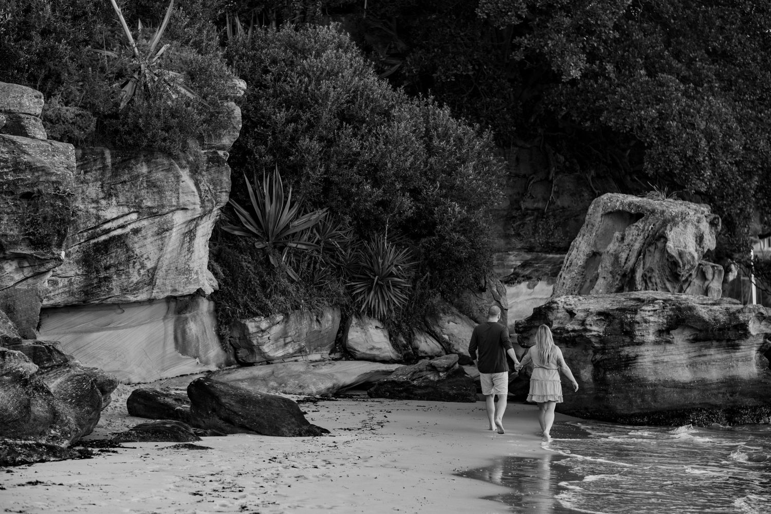 Black and white image of couple walking along rocky beach with cliffs and vegetation in background.