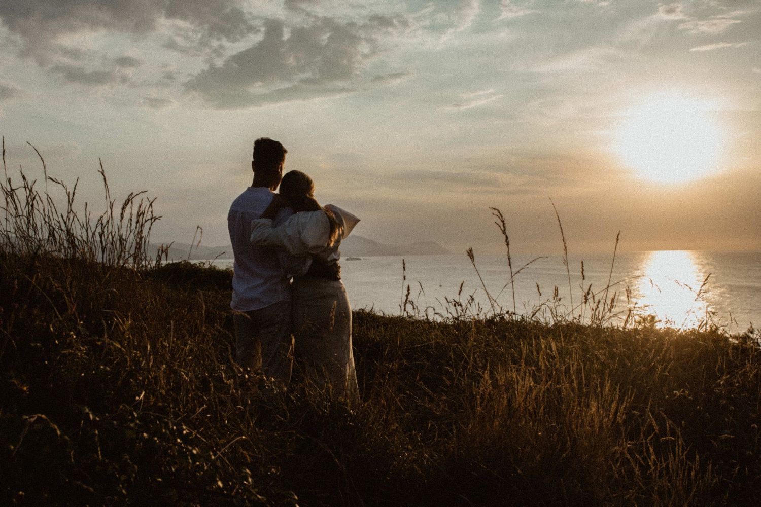 Silhouetted pair watches golden sunset through tall grass on coastal clifftop. Wedding photographer in Mexico Riviera Maya