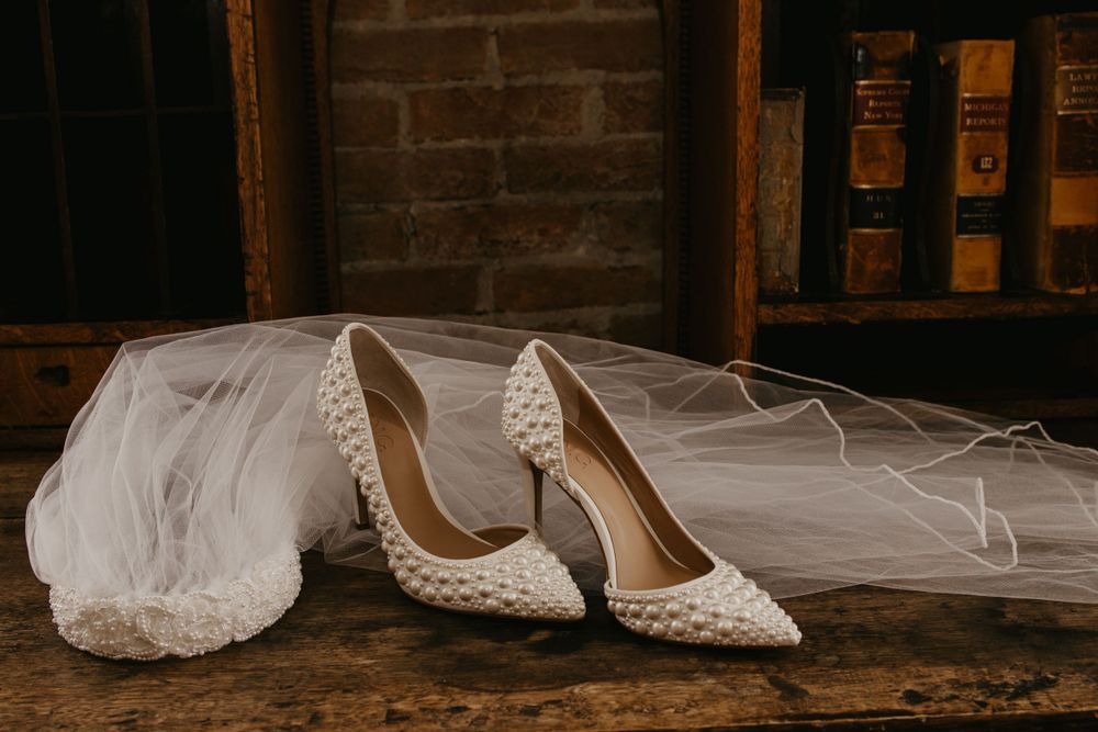Pearl-embellished white bridal heels displayed beside a wedding veil on rustic wooden surface.