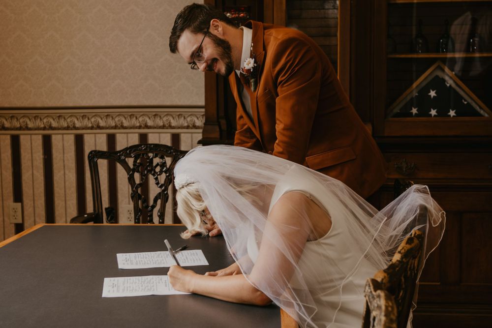 Groom in brown suit watches bride sign marriage certificate at courthouse elopement.