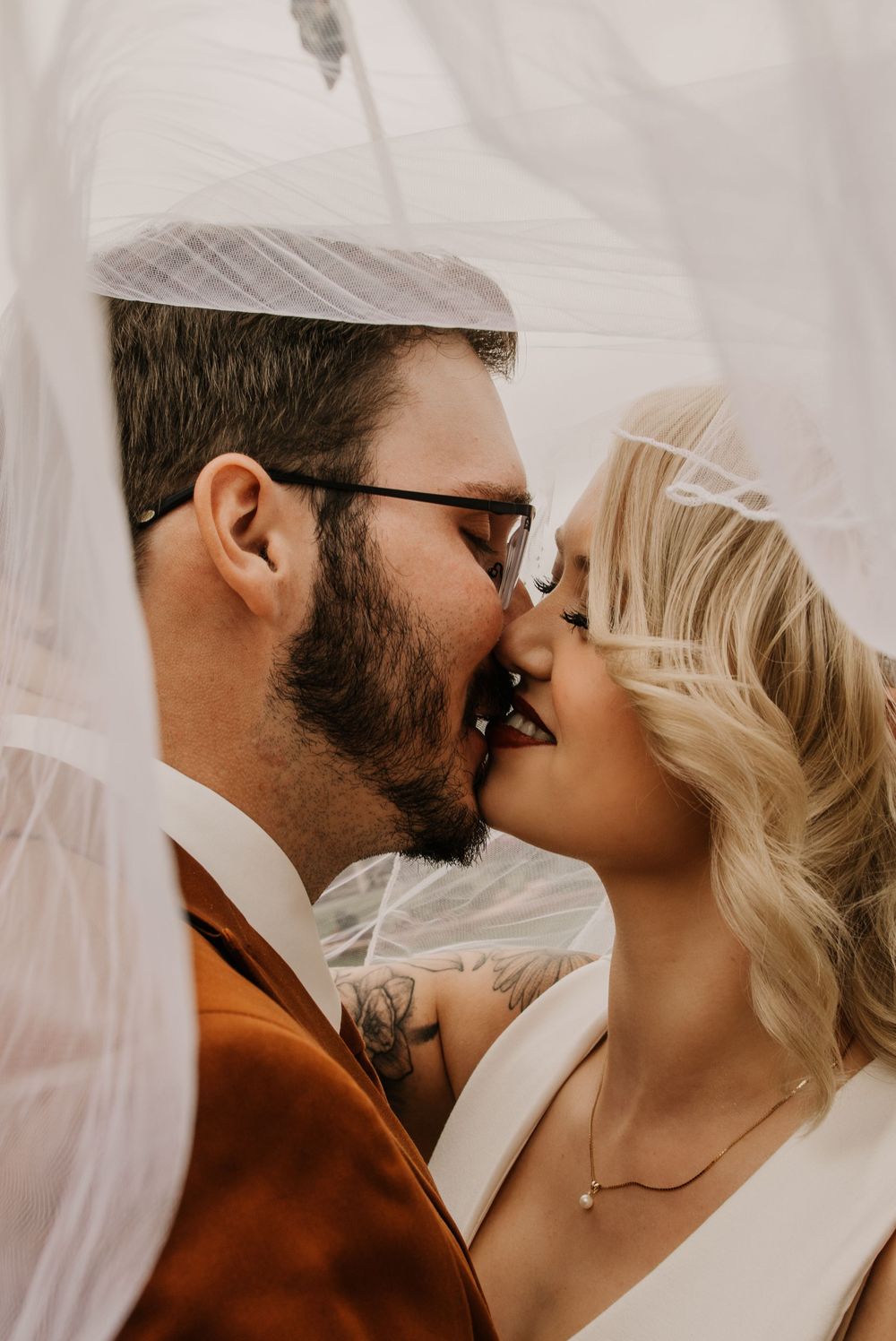 Romantic close-up of bride and groom kissing under flowing white veil, groom wearing glasses and showing tattoos.