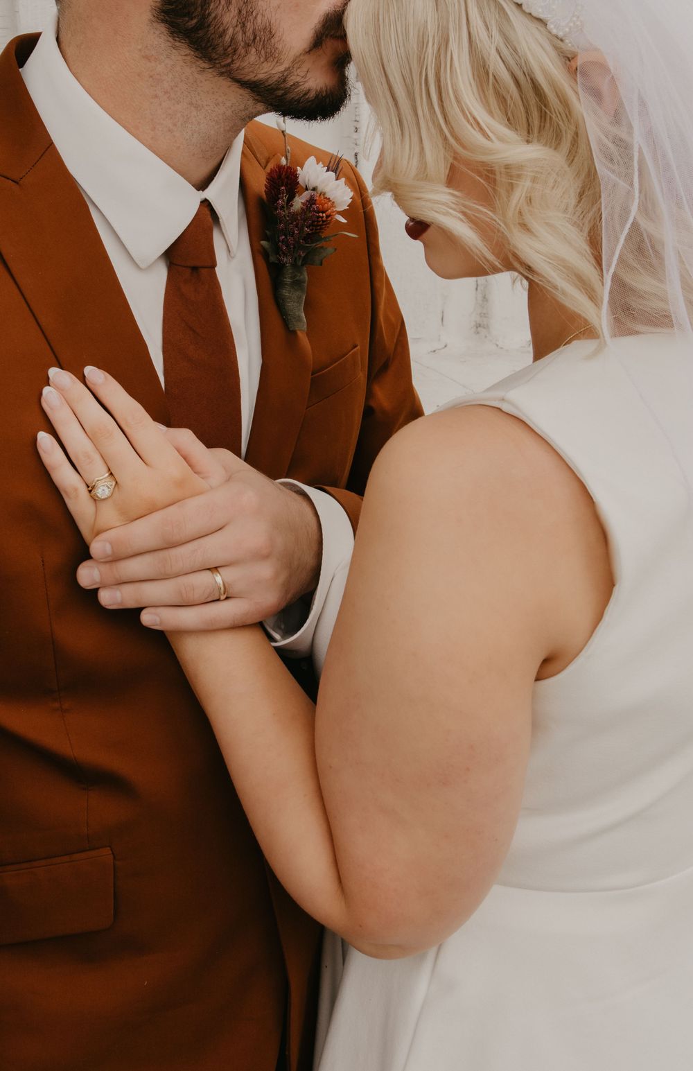 Close-up of bride and groom kissing, groom wearing rust brown suit with floral boutonniere and bride showing wedding ring.