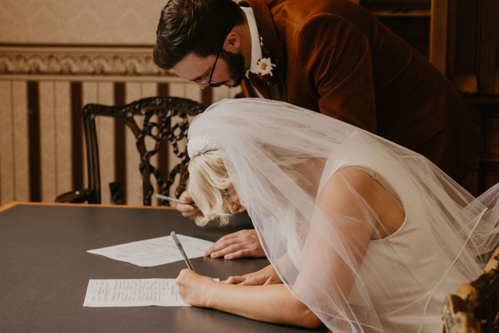 Bride in white mini dress and veil signing wedding documents at wooden table.