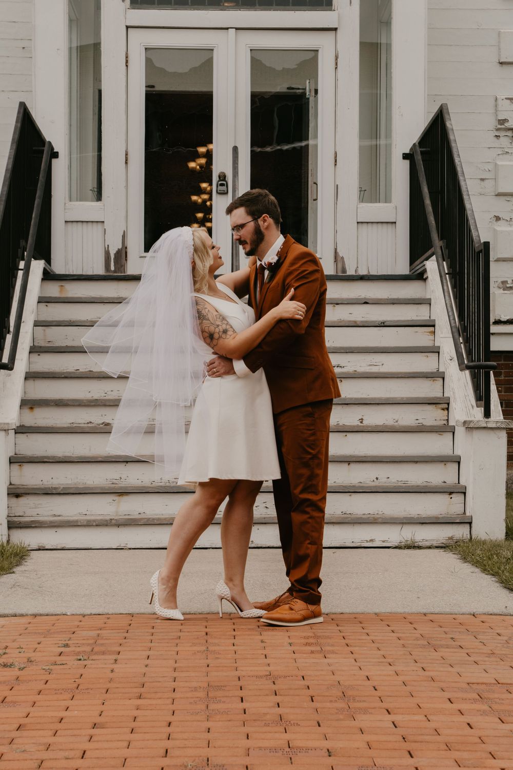 Bride in white mini dress and groom in brown suit embracing outside historic white building.