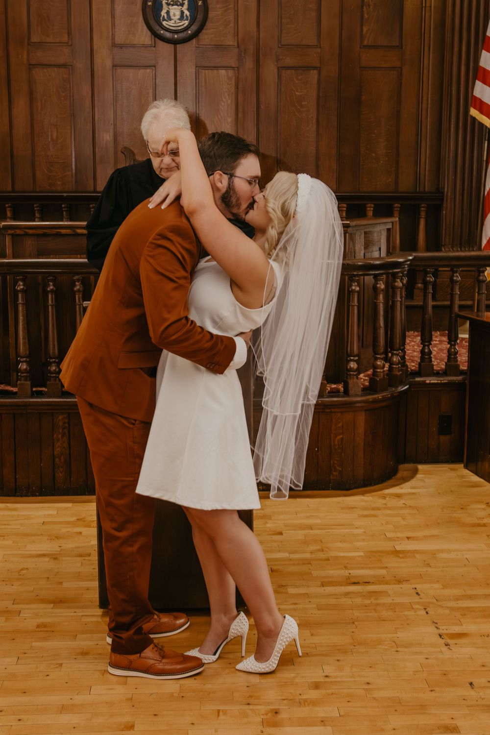 Bride and groom share first kiss in courthouse ceremony with officiant behind them.