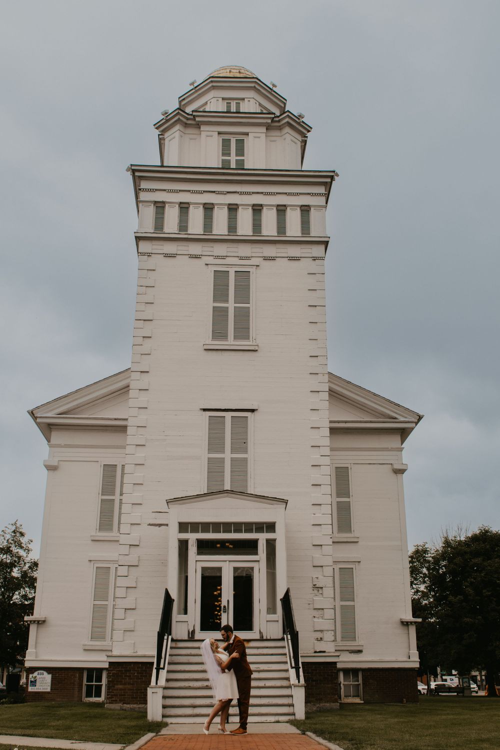 Newlyweds kissing in front of historic white courthouse tower on their wedding day.