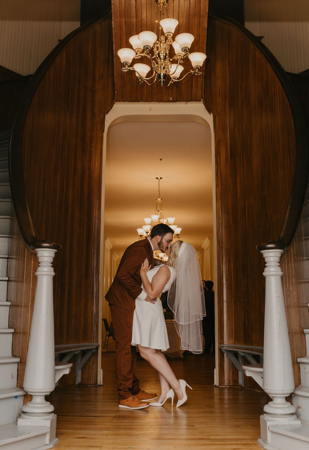 Bride and groom share a kiss in elegant wood-paneled venue doorway beneath a golden chandelier.