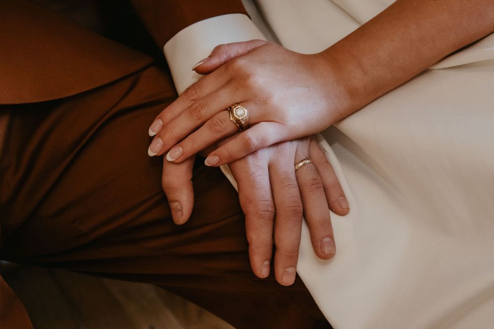 Close-up of newlyweds hands resting together showing gold wedding bands against rust brown suit.