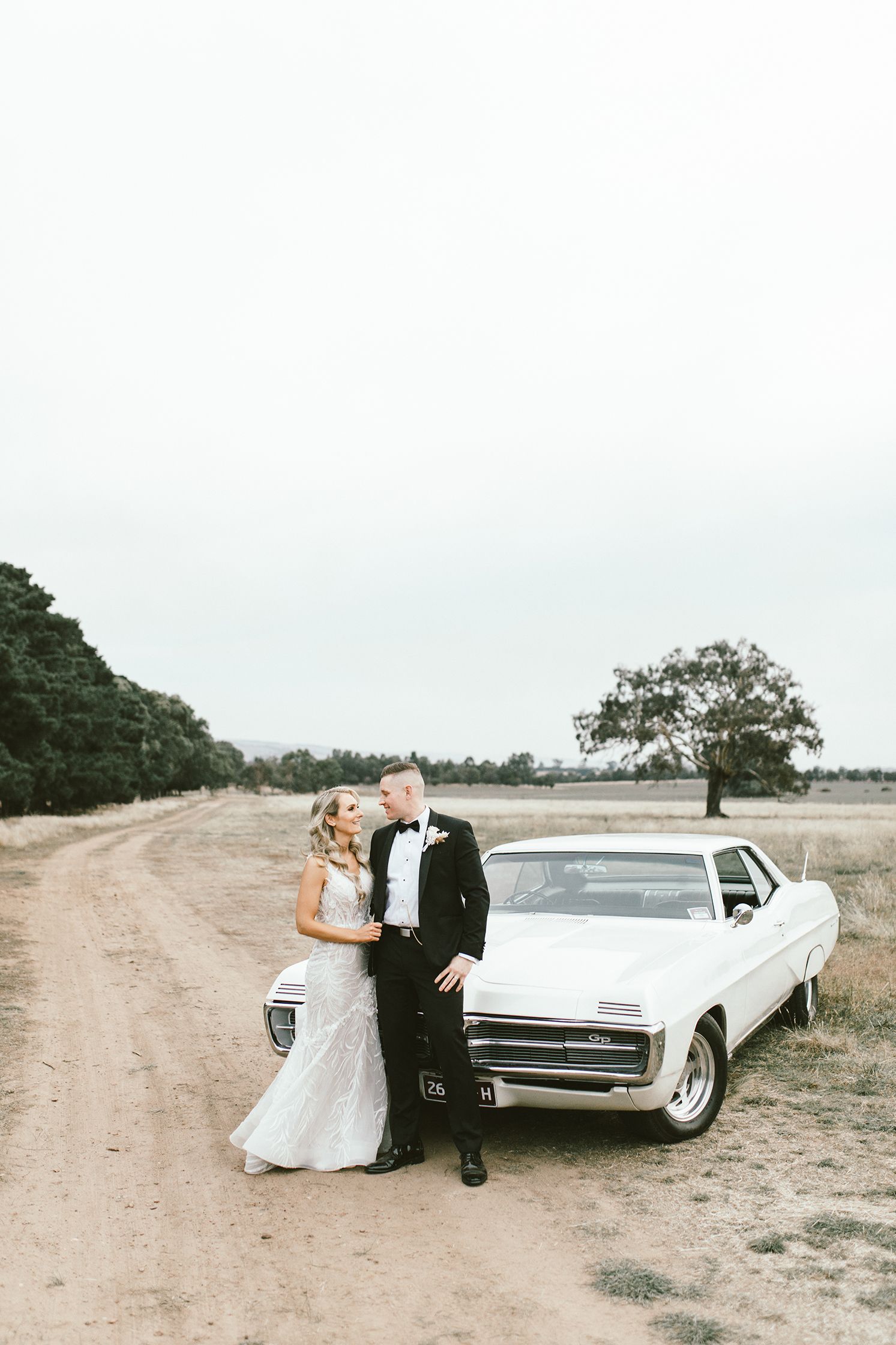 A wedding couple poses alongside a classic white car on a rustic dirt road.