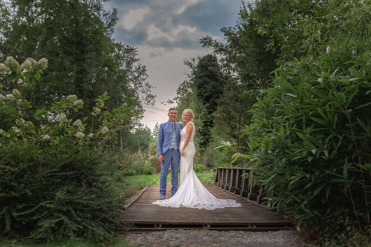 A wedding couple poses on a rustic wooden walkway surrounded by trees at sunset.