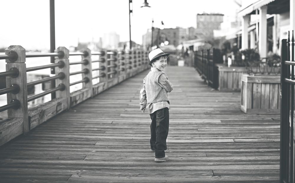 Black and white photo of a small child standing alone on a wooden boardwalk.