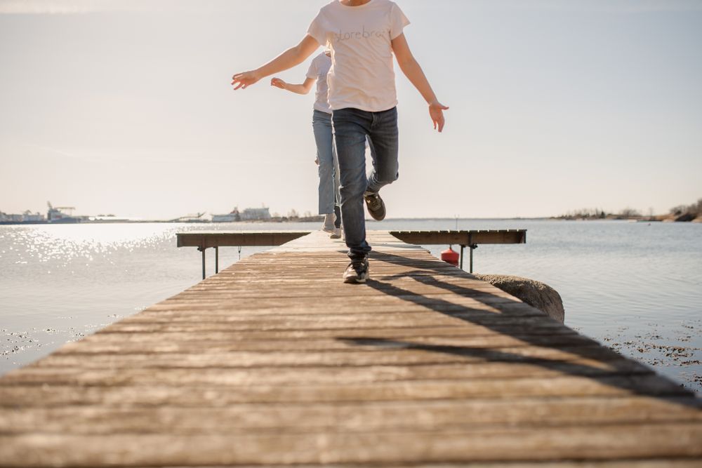 Person walking casually down a wooden dock over tranquil water on a sunny day.