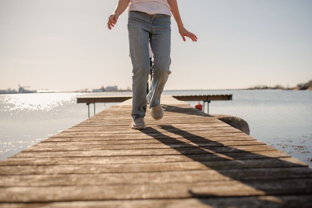 Lower body view of someone walking along a wooden pier over sparkling water.