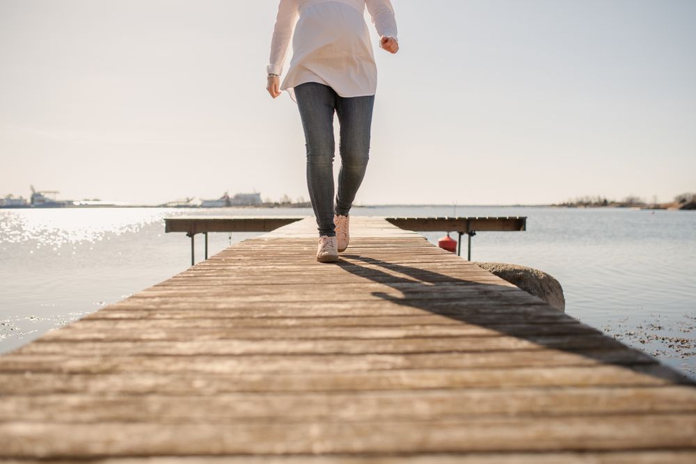 Person in casual attire walking down a sunlit wooden dock over calm waters.