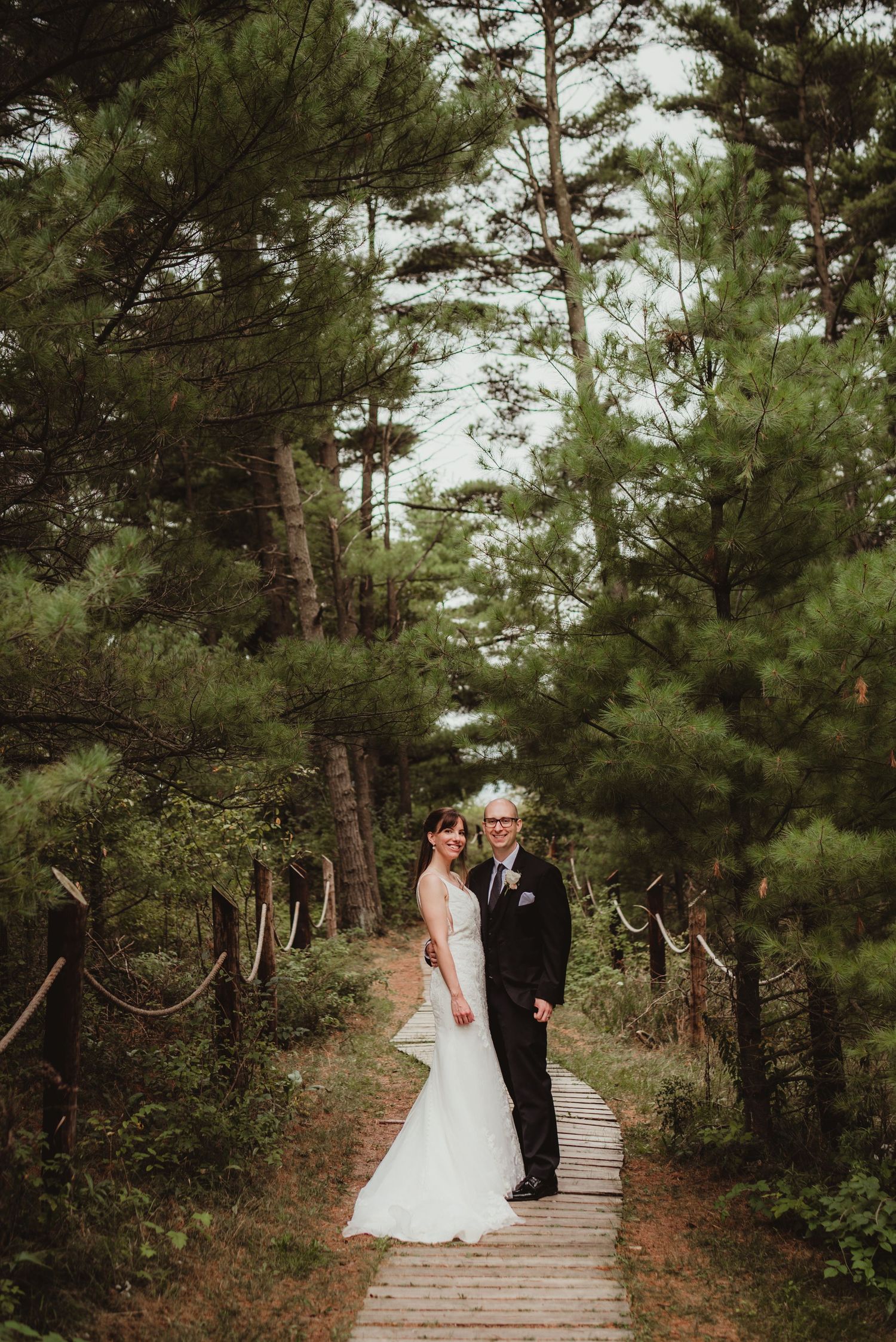 A wedding couple poses on a wooden boardwalk surrounded by tall pine trees in a forest setting.