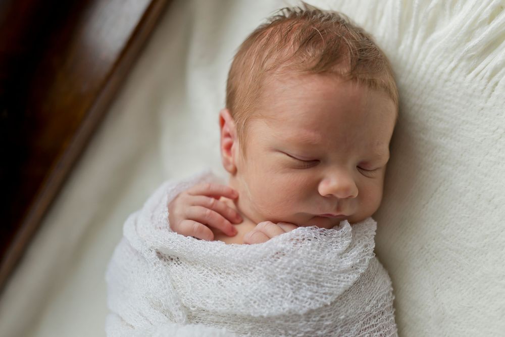 Sleeping newborn wrapped in white swaddle against cream background.