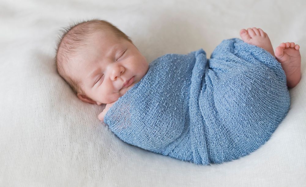Newborn baby wrapped in soft blue swaddle on white background.