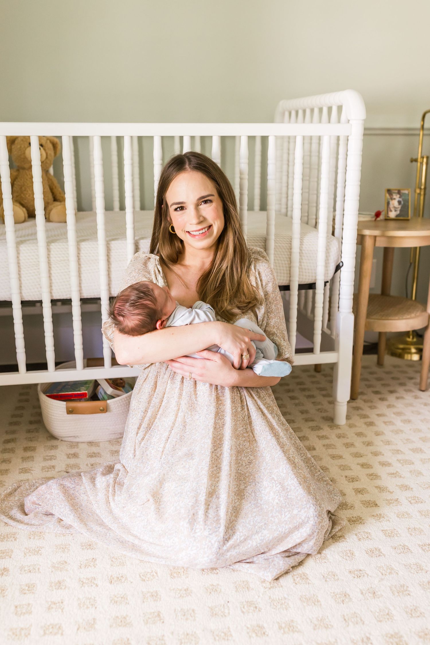 Mother in floral dress sitting on floor beside white crib with newborn.