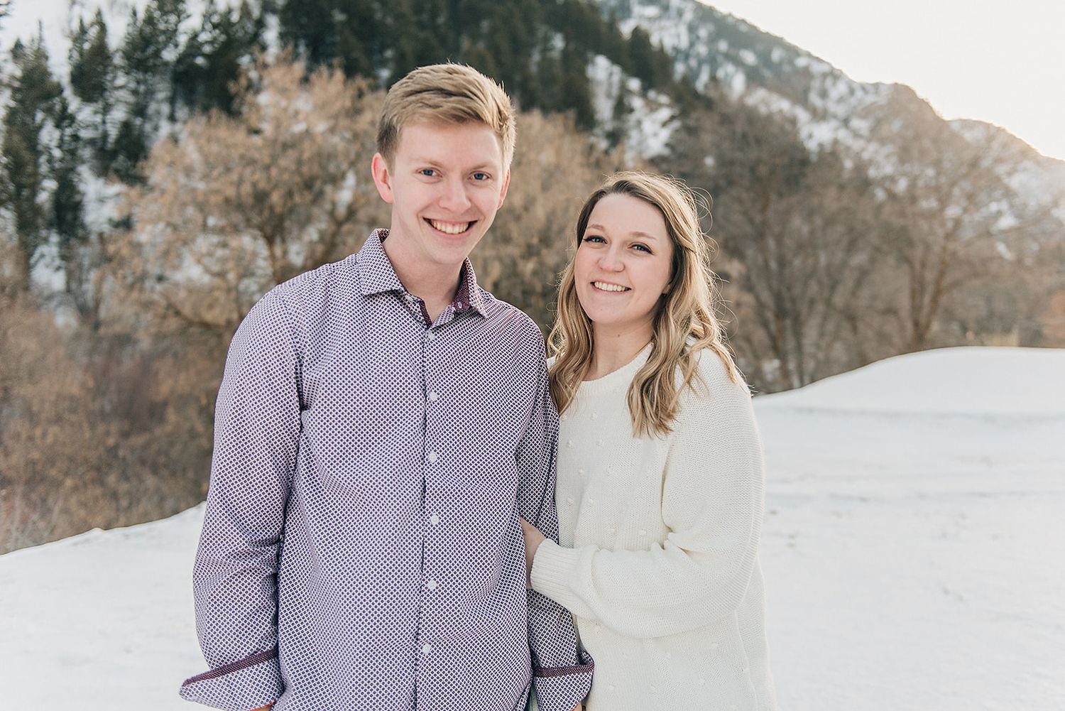 A winter portrait of a couple smiling together with snow-covered mountains in the background.